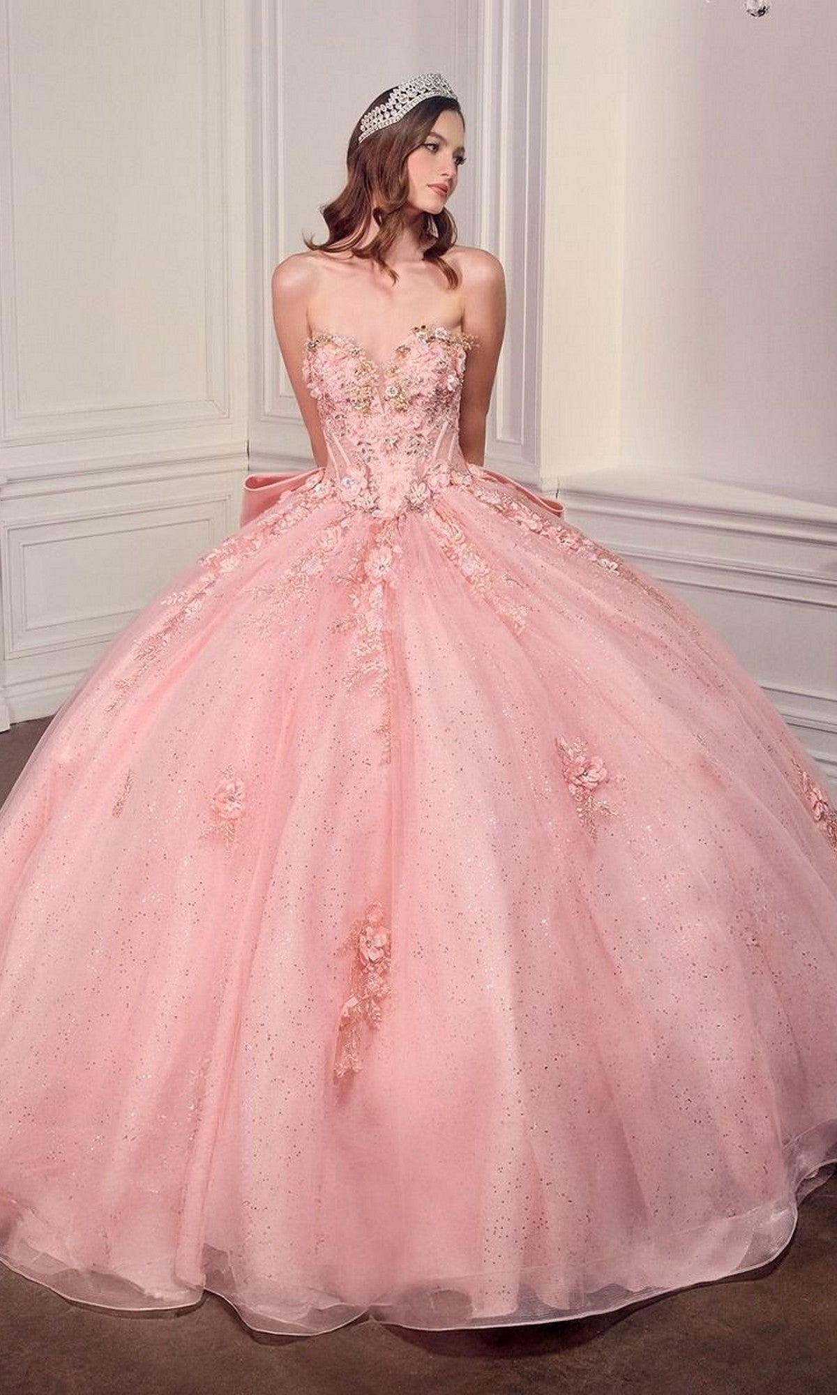 A woman in the Quinceanera Dress 15726 By Ladivine, a light pink strapless gown adorned with 3D flowers and a sparkling tiara, stands in an elegant room with white paneled walls.